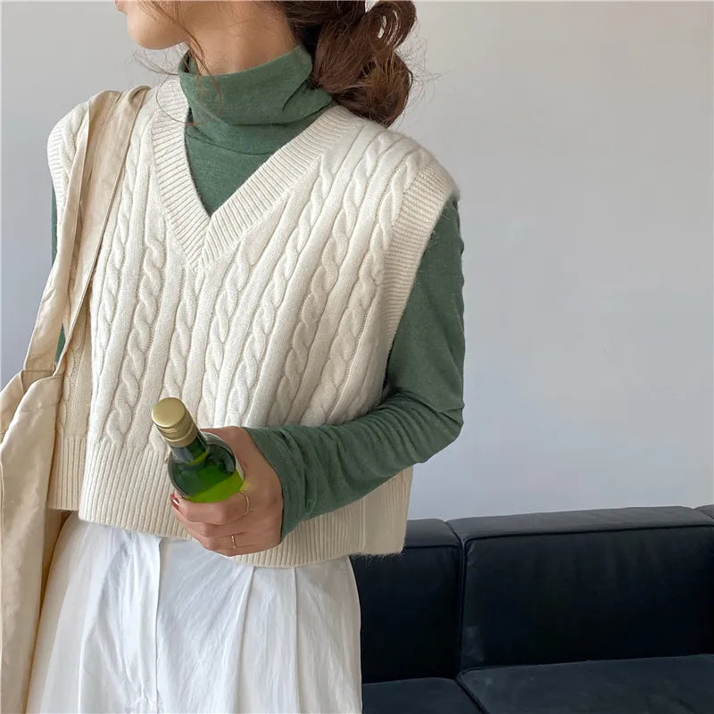 A woman in a cream cable-knit vest and green turtleneck holding a bottle